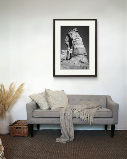 Comfy grey sofa with throw and cushions on top and over it is Framed Fine Art black and white photograph of impressive large arch 
 and visible moon through it. Delicate Arch, Moab, USA, Arches National Park