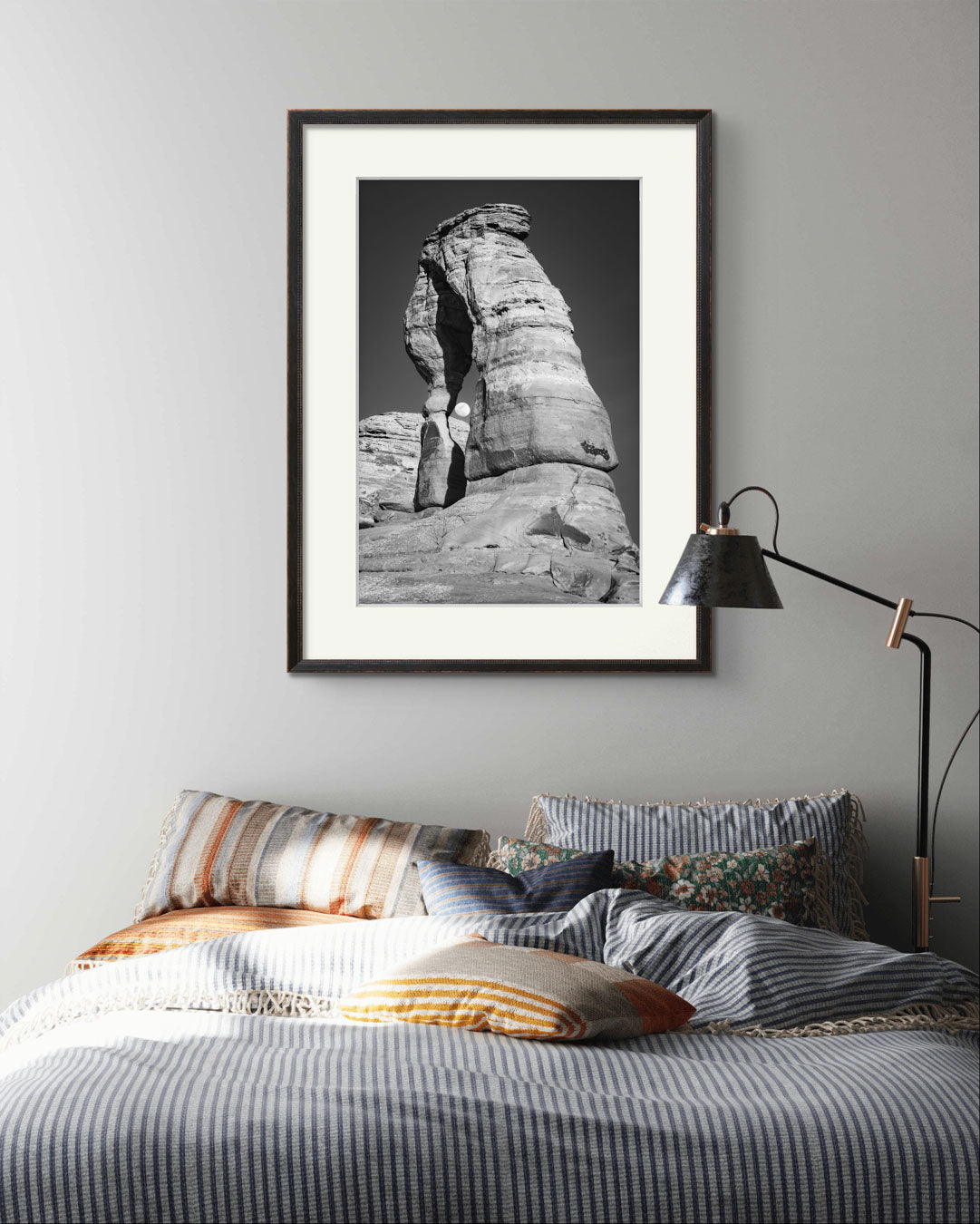 Large bed with stripy bedding on it and over it Framed Fine Art black and white photograph of impressive large arch 
 and visible moon through it. Delicate Arch, Moab, USA, Arches National Park