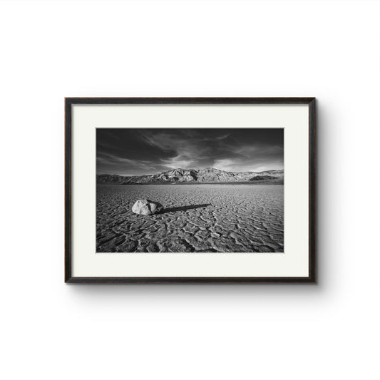 Fine art photography print in black and white. A desert valley in Eastern California, in the northern Mojave Desert. A striking desert landscape in Death Valley National Park. Featuring a solitary rock on the cracked, dry lakebed of the Racetrack Playa, this iconic scene is enhanced by dramatic skies and rugged mountain ranges in the background. Perfect for home decor, wall art, or photography enthusiasts seeking unique nature and landscape prints, this im