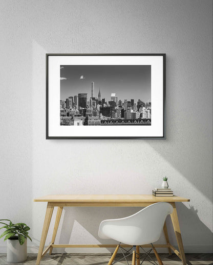 Wooden table by the wall. With white chair next to it. On the wall is  Framed black and white photograph of New York City skyline. All buildings towering behind waterfront. Taken from Brooklyn Bridge side. Manhattan, NYC, USA