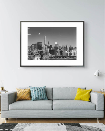 Modern home living room with a large sofa and cushion thrown on it. Over it is hung up Framed black and white photograph of New York City skyline. All buildings towering behind waterfront. Taken from Brooklyn Bridge side. Manhattan, NYC, USA