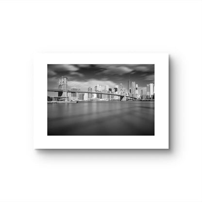 Unframed black and white photograph of Brooklyn Bridge taken from a distance. Dramatic sky and smooth water. NYC. New York City.