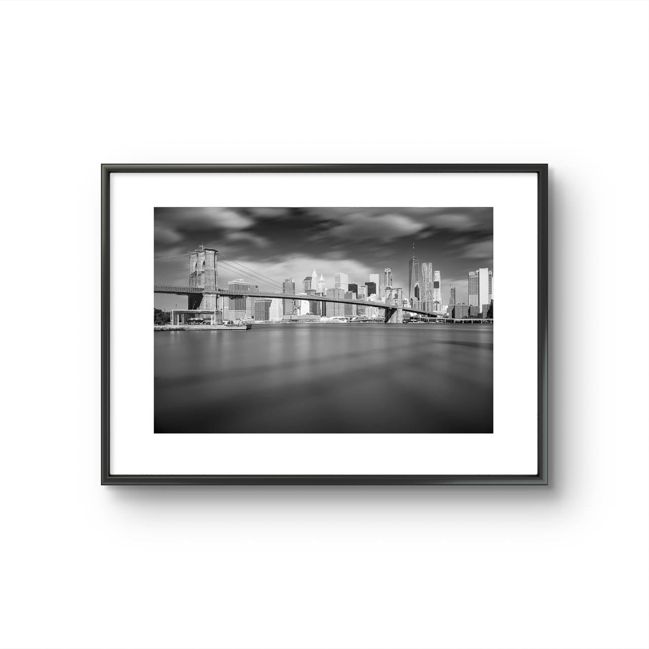 Framed black and white photograph of Brooklyn Bridge taken from a distance. Dramatic sky and smooth water. NYC. New York City.