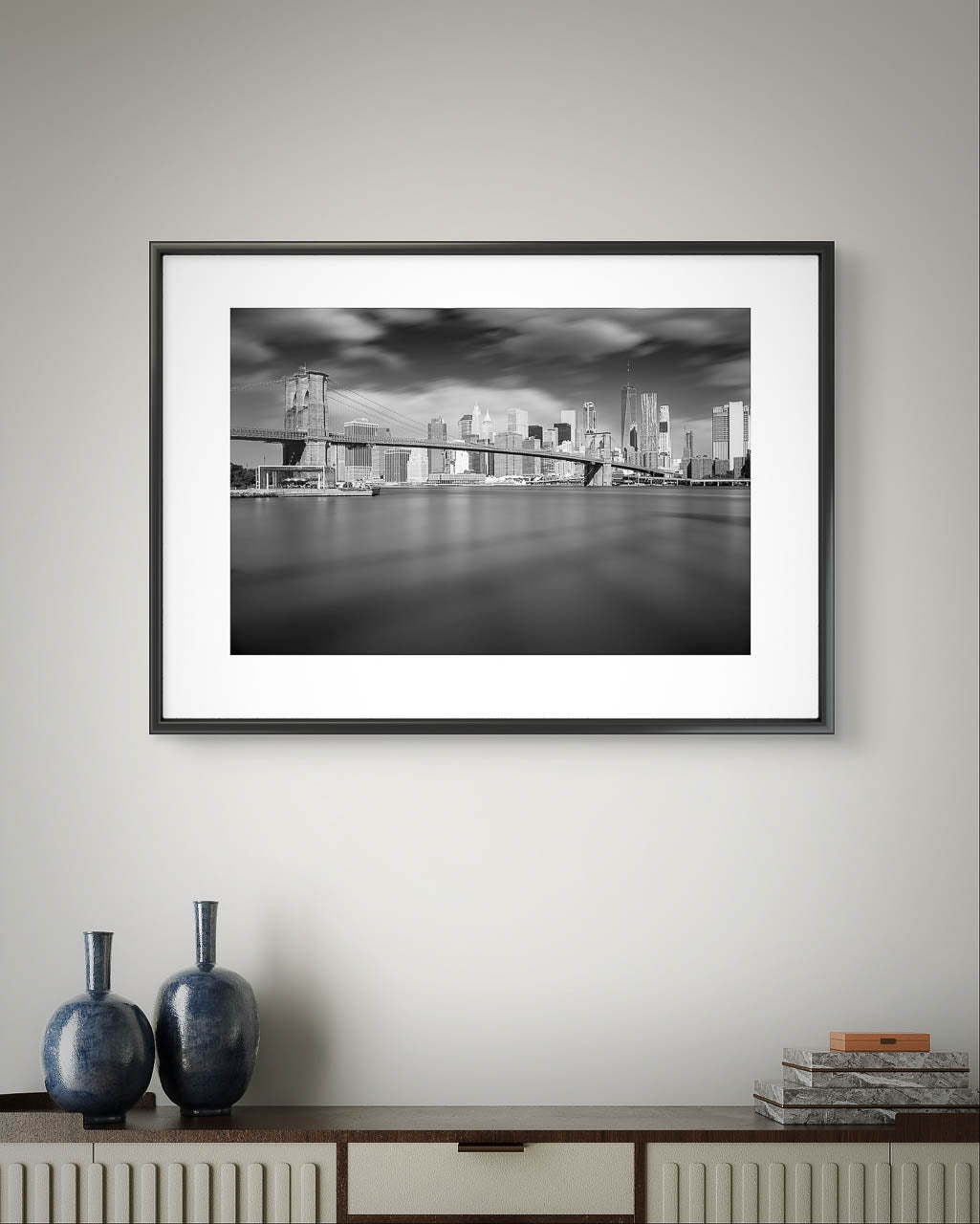 Modern console table with two vases on top. On the wall is a Framed black and white photograph of Brooklyn Bridge taken from a distance. Dramatic sky and smooth water. NYC. New York City.