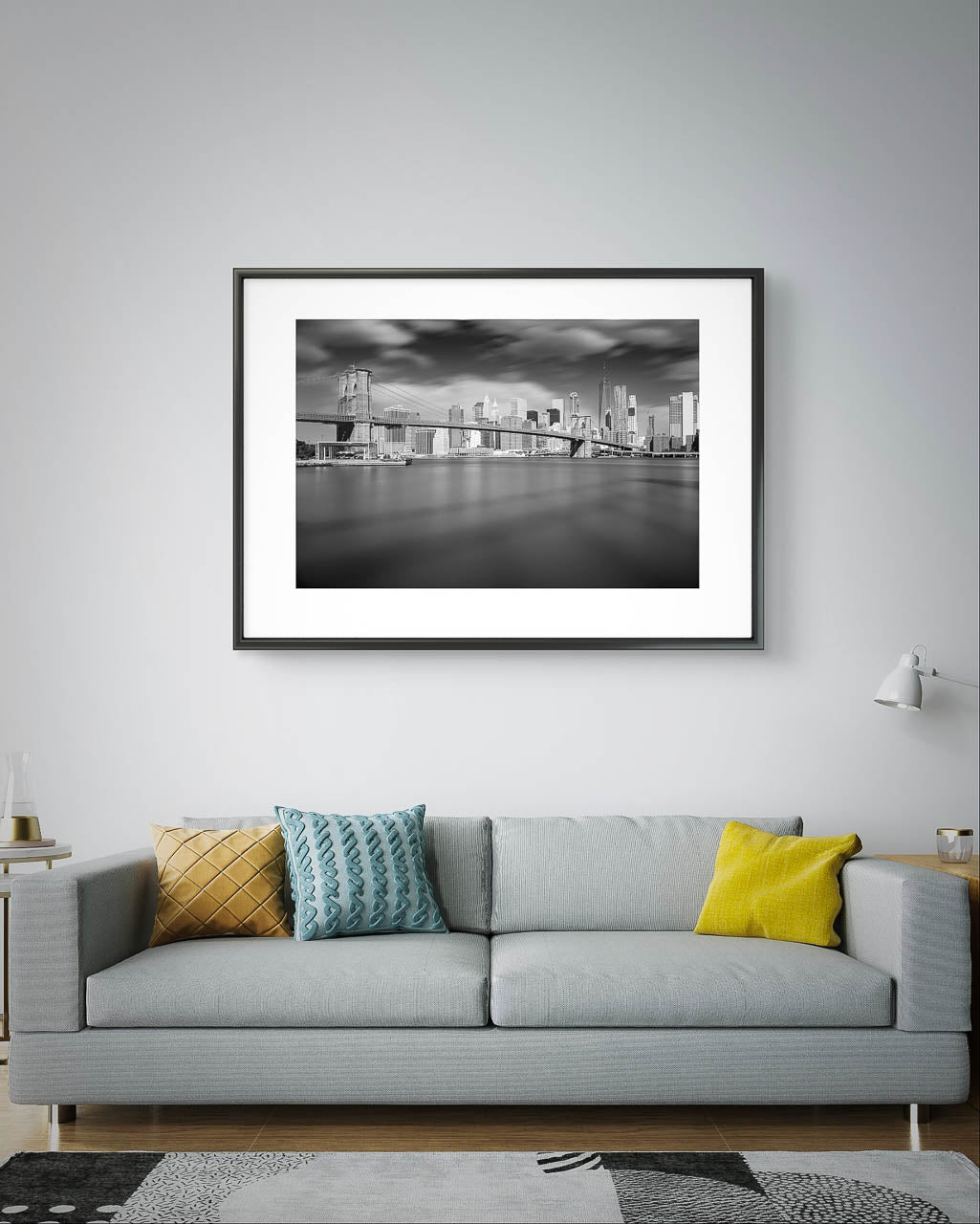 Large grey sofa with colorful cushions . Over it Framed black and white photograph of Brooklyn Bridge taken from a distance. Dramatic sky and smooth water. NYC. New York City.