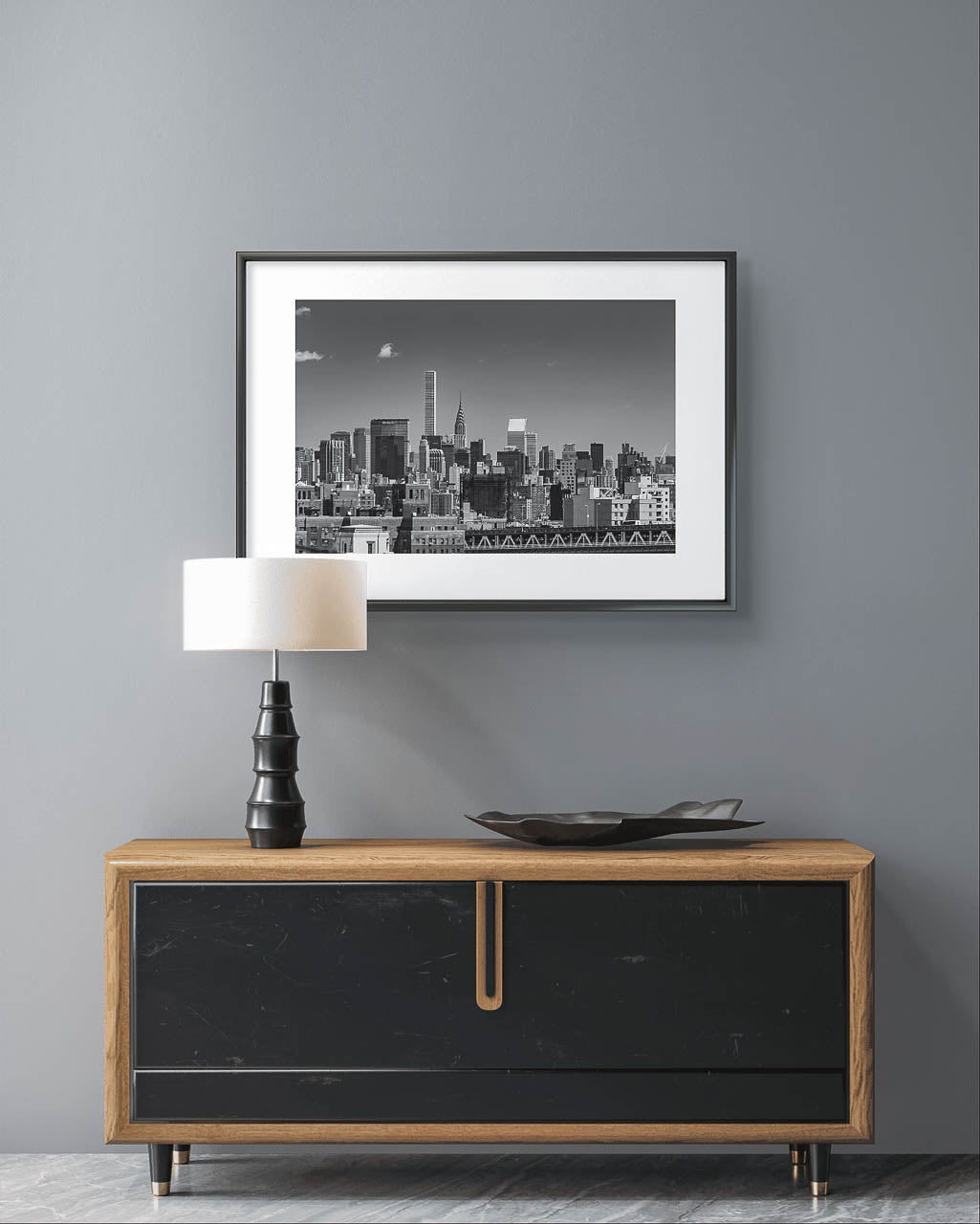 Over a modern console table on the grey wall is Framed black and white photograph of New York City skyline. All buildings towering behind waterfront. Taken from Brooklyn Bridge side. Manhattan, NYC, USA