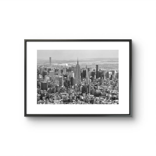 FRamed black and white fine art photography print. Aerial view of Manhattan with the iconic Empire State Building reigning supreme at its center. The photograph captures the city in all its grandeur, with skyscrapers reaching for the sky and the bustling streets below. NYC. New York City